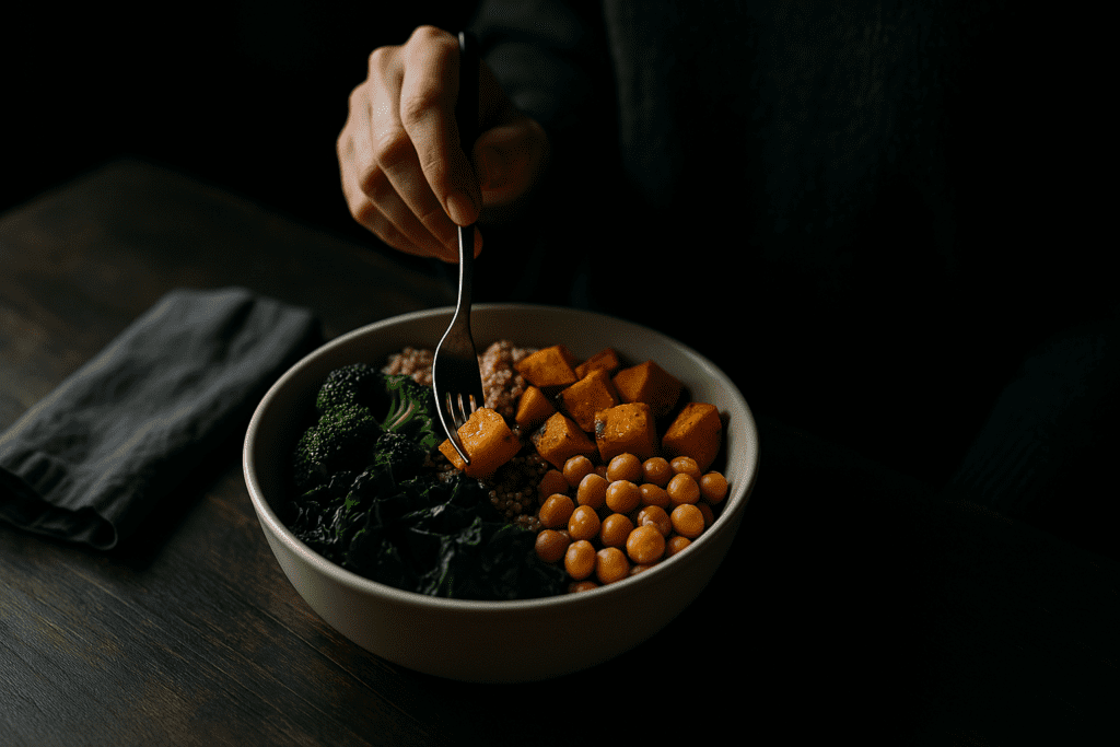 Healthy Plant-Based Dinners Made Easy: Best Whole Food Plant-Based Recipes for Beginners and Beyond 3 A close-up shot shows a hand reaching in with a fork to enjoy a colorful plant-based dinner bowl in moody, dramatic lighting on a dark wooden table. The vibrant whole food ingredients—broccoli, kale, chickpeas, quinoa, and roasted sweet potatoes—stand out against the dim background, capturing the richness and simplicity of healthy plant-based dinners made easy.