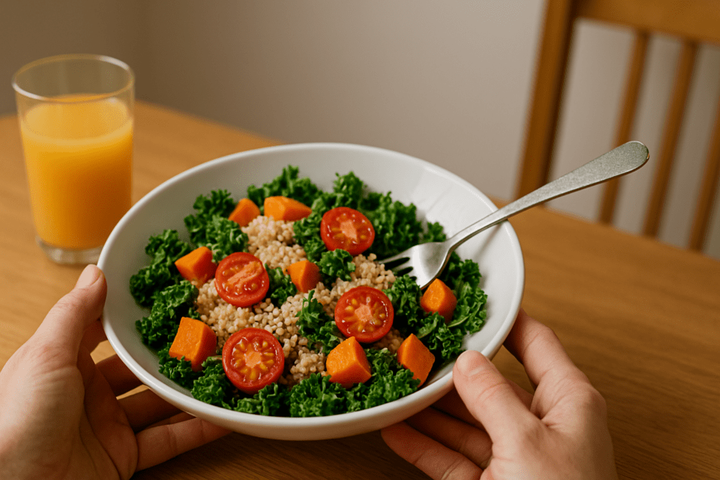 A pair of fair-skinned hands holding a white plate filled with kale, quinoa, cherry tomatoes, and roasted sweet potatoes, placed on a wooden table with a glass of juice in the background.